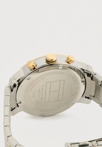 Tommy Hilfiger rozsdamentes acél karóra hátlapja, aranyszínű nyomógombokkal és gravírozott márkanevével, valamint specifikációkkal.