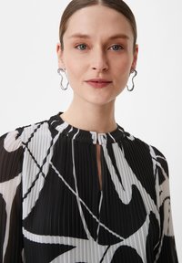 Czarna i biała plisowana bluzka z okrągłym dekoltem, z abstrakcyjnymi wzorami i detalem w postaci klucza z przodu. Modelka nosi srebrne kolczyki w kształcie abstrakcyjnym.