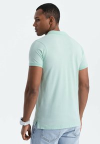 Smaragdittut kjøns skjorte med korte ermer, fin tekstur, sidesplitt, parret med lyseblå denimjeans, sett fra baksiden.