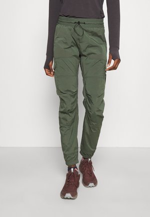 Personne portant un pantalon cargo vert foncé avec des poignets élastiques et des chaussures de sport marron, debout devant un fond uni.