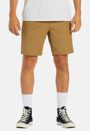 Hombre con pantalones cortos marrón claro hasta la rodilla, calcetines blancos, zapatillas negras altas y camisa blanca, de pie contra un fondo liso.