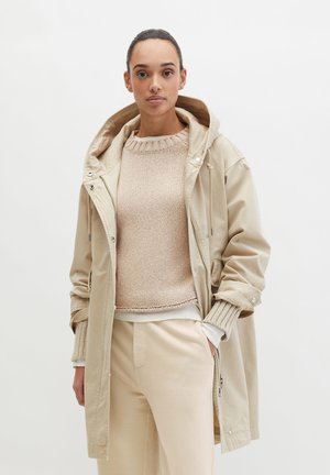 Femme portant une veste à capuche beige par-dessus un pull léger en maille et un pantalon de couleur crème, debout devant un fond blanc uni.