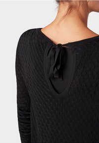 Frau, die einen strukturierten schwarzen Pullover mit einem Schlüssel-Ausschnitt auf dem Rücken trägt, der mit einer Stoffschleife im Nacken gebunden ist.