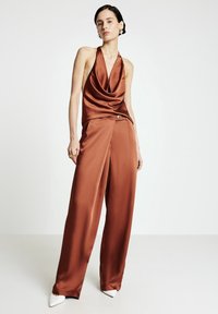 Braunes Satin-Halfter-Top mit drapiertem Ausschnitt, kombiniert mit weit geschnittenen braunen Hosen. Auffälliges Knopfdetail an der Taille, abgerundet mit weißen High Heels.