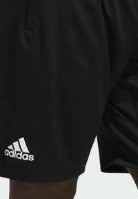 Mustat urheilushortsit, jotka on valmistettu kevyestä kankaasta, sisältävät joustavan vyötärön ja sivutaskut. Valkoinen Adidas-logo on näkyvissä helman kohdalla.