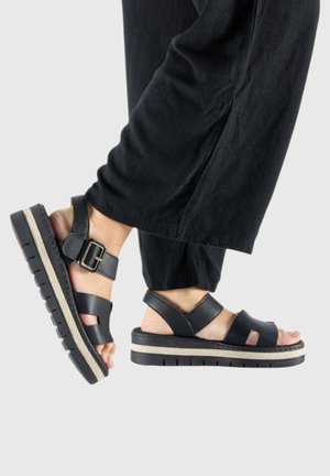 Pieds portant des sandales à plateforme noires avec des brides et des boucles à la cheville, associés à un pantalon noir ample sur un fond uni.