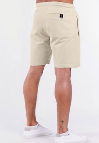 Beige Baumwollshorts mit einem elastischen Bund, seitlichen Taschen und einer Gesäßtasche, die mit einem schwarzen Logo-Patch versehen ist. Glatte Textur, lockere Passform.
