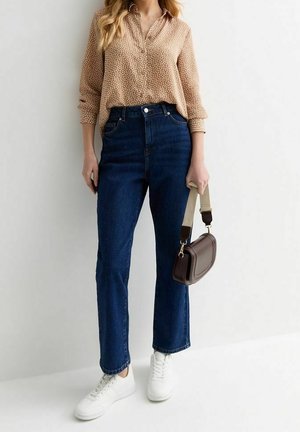Femme portant un chemisier beige à motifs, un jean bleu foncé taille haute, des baskets blanches, tenant un petit sac à main marron avec une bandoulière beige.