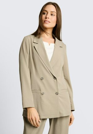 Vrouw draagt een beige double-breasted blazer met inkeping revers over een wit topje, staand tegen een effen lichte achtergrond.