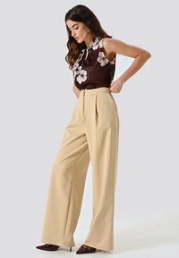 Pantaloni plissettati beige chiaro e una blusa di chiffon floreale scura con dettaglio annodato al collo; texture liscia, design a gamba larga.