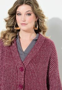 Burgunderfärgad stickad cardigan med V-ringning, två lila knappar och texturerade detaljer. Bärs över en grå topp med en mjuk finish.