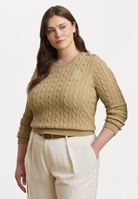 Cable-knit beige genser med rund hals, som har en liten brodert logo og knappdetaljer på skulderen. Parret med lyse bukser.