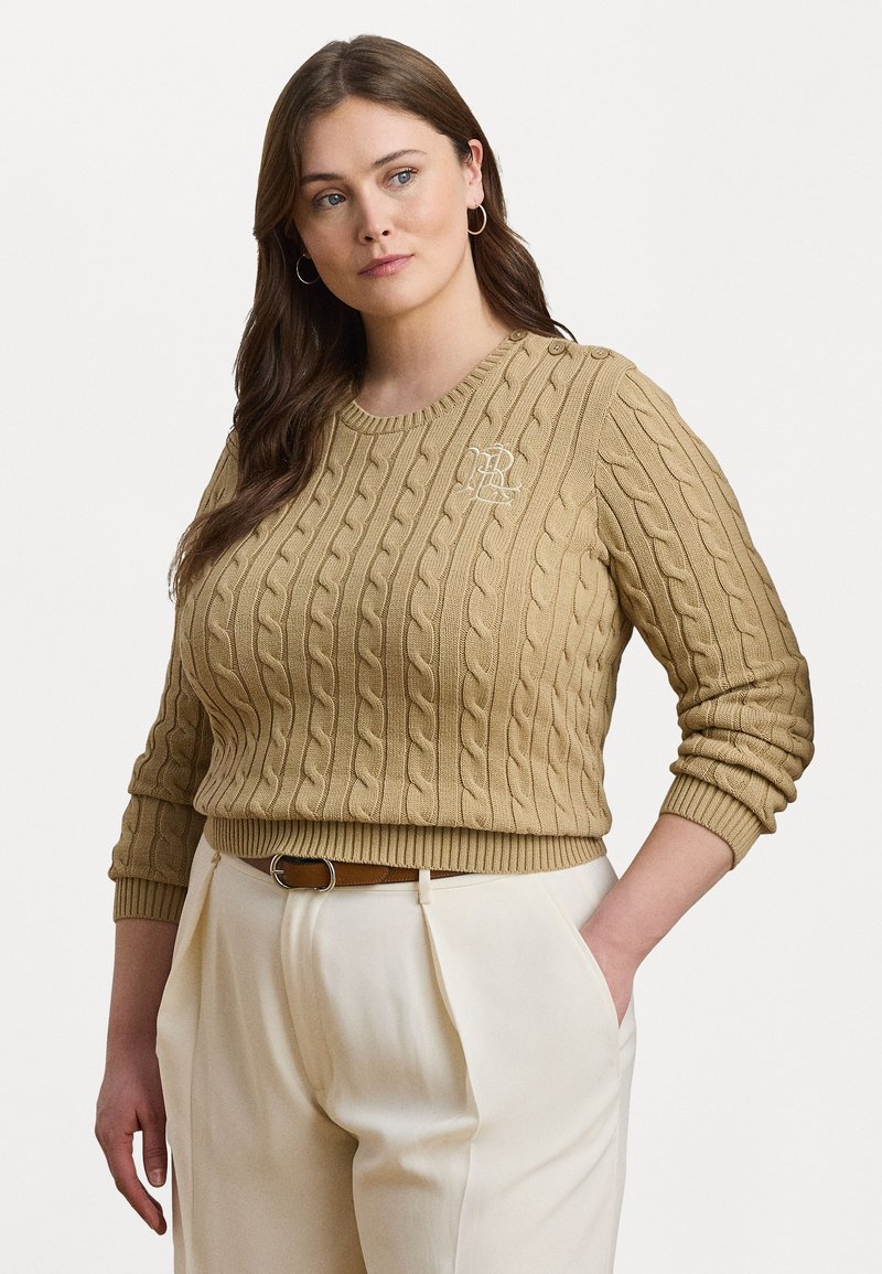Cable-knit beige genser med rund hals, som har en liten brodert logo og knappdetaljer på skulderen. Parret med lyse bukser.