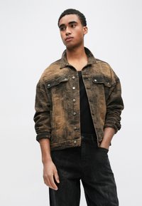 Brūna valkāta džinsa jaka ar pogām priekšpusē, divām krūšu kabatām un satītām piedurknēm. Valkāta virs melnas blūzes, kombinēta ar tumšām biksēm.