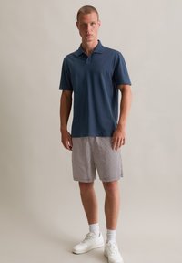 Marineblauwe polo met korte mouwen, gecombineerd met geruite shorts in wit en rood. Witte sneakers maken de outfit compleet. Neutrale achtergrond.