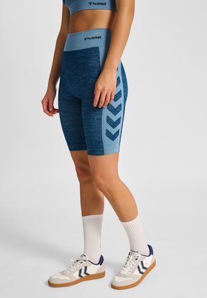Blaue, hoch taillierte Shorts aus strukturiertem Stoff, mit einem kontrastierenden Seitenpaneel mit Chevron-Mustern, kombiniert mit weißen Sneakers und Socken.