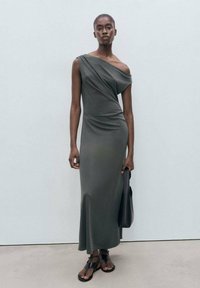 Long, one-shoulder, dark green dress made from soft fabric, featuring draped design and a slightly flared hem. Worn with black sandals and a bag.