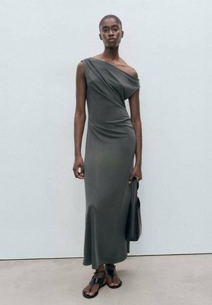 Robe longue, à une épaule, de couleur vert foncé, fabriquée dans un tissu doux, avec un design drapé et un ourlet légèrement évasé. Portée avec des sandales noires et un sac.