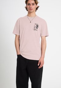 Jeune homme portant un t-shirt rose pâle avec des masques de comédie et de tragédie en noir et blanc et le logo "Volcom Stone Co.", associé à un pantalon noir.