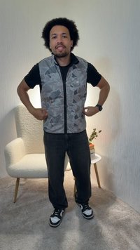 Uomo con capelli ricci che indossa una camicia nera, un gilet grigio a fantasia, pantaloni scuri e sneakers, in piedi all'interno vicino a una poltrona bianca e un piccolo tavolo con dei fiori.