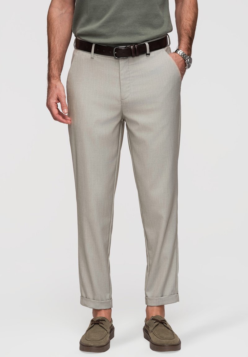 Homme portant un pantalon gris clair sur mesure avec revers, une ceinture marron, des chaussures vert olive, un t-shirt vert et une montre argentée.