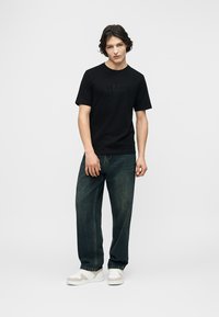 Νέος άντρας όρθιος, φορώντας μαύρο κοντομάνικο μπλουζάκι, σκούρο φαρδύ τζιν και λευκά αθλητικά παπούτσια, μπροστά σε απλό φωτεινό φόντο.