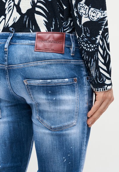Κοντινό πλάνο ατόμου που φορά ξεθωριασμένο μπλε τζιν με καφέ δερμάτινο ταμπελάκι DSQUARED2 και μαύρη και λευκή πουλόβερ με μοτίβο.