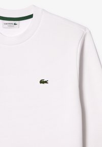 Weißer Sweatshirt mit rundem Halsausschnitt, grünem Kragen und kleinem gesticktem grünen Krokodil-Logo auf der linken Brust. Klassische Passform.