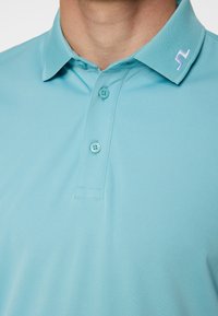 Homme portant un polo bleu clair avec deux boutons fermés et un logo blanc brodé sur le col.