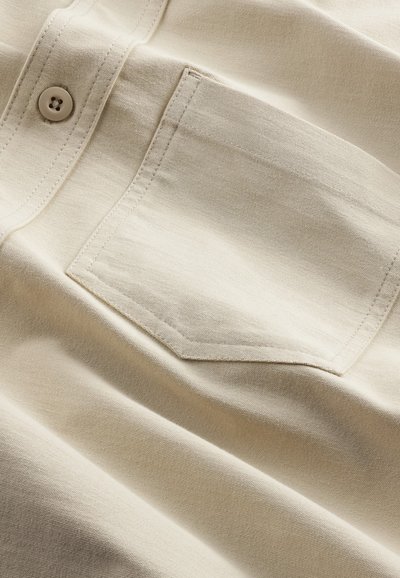 Beige bomullsskjorta med en bröstficka och knapp. Tyget har en slät textur med subtil glans och sydda detaljer.