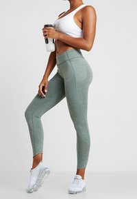 Grågröna högmidjade leggings gjorda av stretchigt material, med perforerade detaljer nära anklarna, kombinerade med en vit sport-bh och transparenta träningsskor.