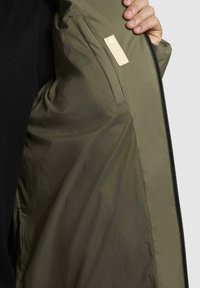 Oliven grøn jakke indvendigt med glat tekstur og et stofmærke. Har en lynlåslukning og en let løs, afslappet pasform.