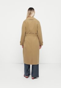 Trench coat lungo con cintura in un tessuto marrone chiaro. Presenta un colletto classico e dettagli sui polsini. Vista posteriore con taglio dritto e minimalismo nei dettagli.