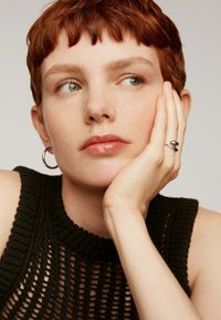 Top sin mangas de punto negro con un patrón acanalado; la modelo tiene el cabello rojo corto, lleva un pendiente de aro plateado y un anillo plateado con dos acentos redondos.