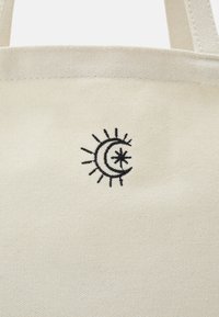 Sac cabas en toile beige avec un symbole de soleil, lune et étoile brodé en noir près du centre supérieur. Deux poignées sont partiellement visibles.
