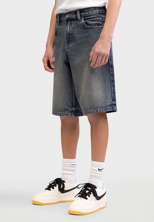 Osoba nosiaca vybledlé modré rifľové šortky, biele ponožky Nike a biele a žlté tenisky s čiernymi šnúrkami a logom Swoosh.