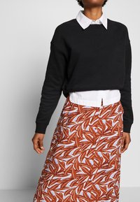 Felpa nera cropped con spalle abbassate indossata sopra una camicia bianca con colletto. La gonna presenta un motivo di foglie marroni su uno sfondo chiaro.