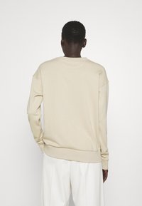 Beżowy sweter z prążkowanymi mankietami i dołem, z okrągłym dekoltem i opuszczonymi ramionami, zestawiony z białymi spodniami o szerokich nogawkach.