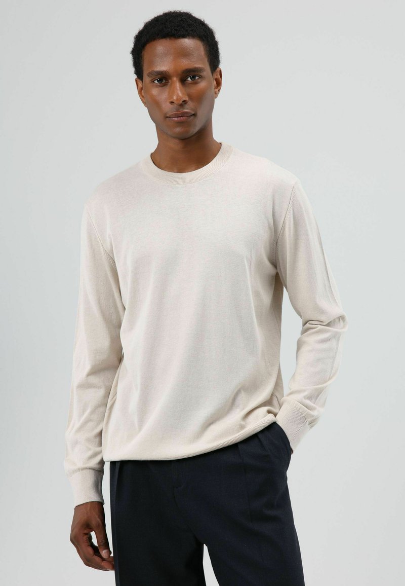 Lichtbeige longsleeve T-shirt gemaakt van zachte stof. Voorzien van een ronde halslijn en geribbelde manchetten, gecombineerd met een donkere broek.