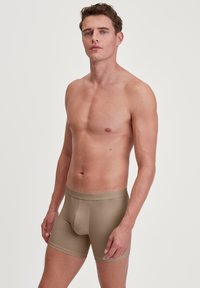 Boxer in beige con design aderente, realizzati in tessuto morbido. Presentano un elastico soffice in vita. Modello mostrato in posizione eretta, torace nudo.