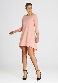 Rosa asymmetrisches Kleid mit rundem Ausschnitt, drei Viertel Ärmel und glatter Textur. Schwarze High-Heels vervollständigen den Look.