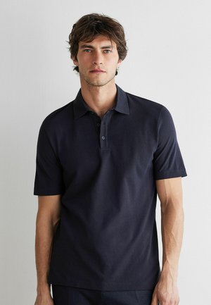 Junger Mann mit zerzausten braunen Haaren, der ein dunkelblaues Poloshirt mit kurzen Ärmeln vor einem schlichten weißen Hintergrund trägt.