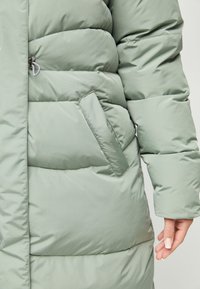 Vihreä puffer-takki, jossa on vetoketjullinen tasku, tikattu muotoilu ja sileä, kiiltävä pinta, joka myötäilee vartalon muotoja.