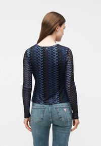 Γυναίκα με μακριά καστανά μαλλιά που φοράει μπλε και μαύρο μακρυμάνικο μπλουζάκι με σχέδιο και ανοιχτό μπλε τζιν, γυρισμένη προς τα πίσω μπροστά σε απλό φόντο.