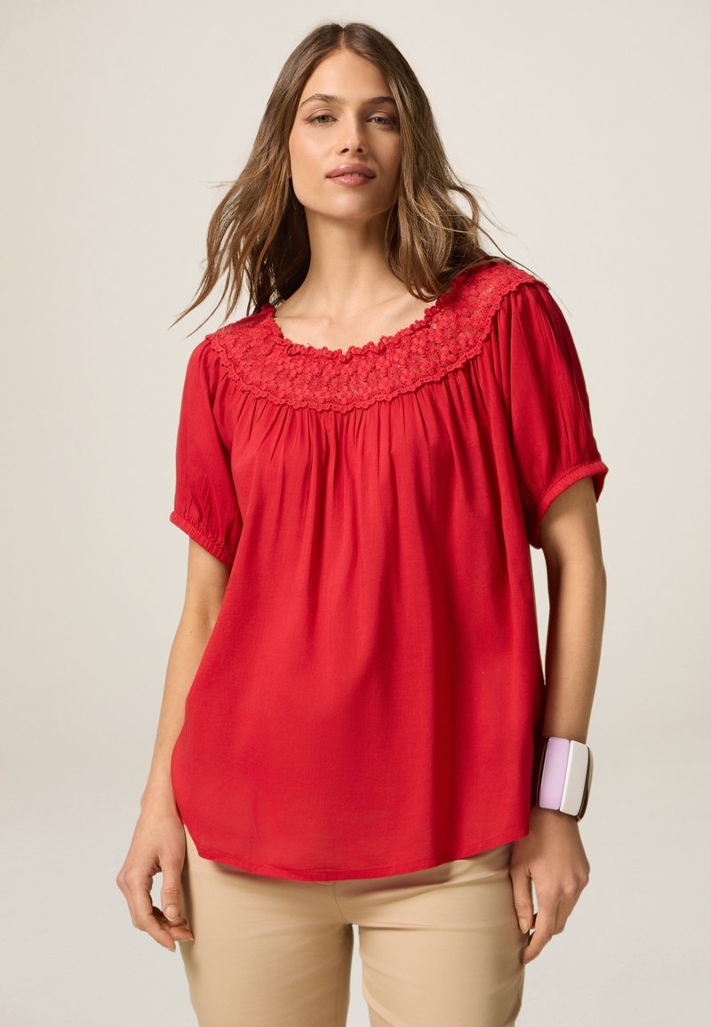 Blusa roja con un escote bordado en forma de ondas, mangas cortas abullonadas y un corte relajado, hecha de un tejido ligero. Combinada con pantalones beiges.