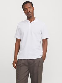 Polo shirt en coton blanc à manches courtes, avec une patte de boutonnage et un logo discret sur la poitrine, associé à un pantalon sombre à plis.