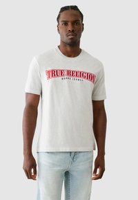 Pilka trumpa rankovėmis palaidinė su raudonu ir mėlynu "TRUE RELIGION" logotipu per krūtinę, derinama su šviesiai mėlynomis nusidėvėjusiomis džinsinėmis kelnėmis.