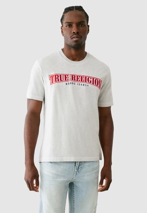 Szürke rövid ujjú póló, amelyen piros és kék "TRUE RELIGION" logo látható a mellkasnál, világoskék megkoptatott farmernadrággal párosítva.