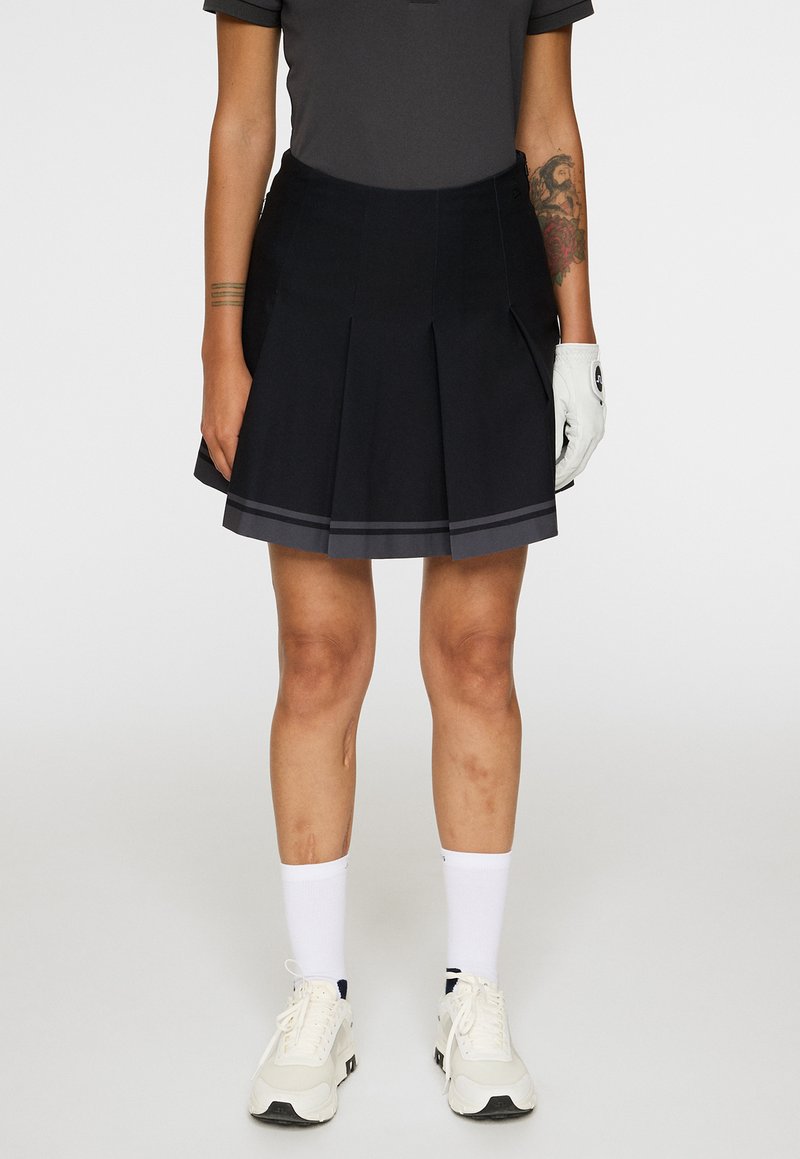 Persoon in een zwarte geplooide rok, donker shirt, witte sok sneakers, witte golfhandschoen en zichtbare tatoeages op de armen, staand tegen een effen achtergrond.