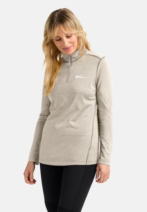 Frau mit langen blonden Haaren, die einen hellgrauen Reißverschluss-Fleece-Pullover und schwarze Leggings trägt, vor einem einfarbigen Hintergrund steht.